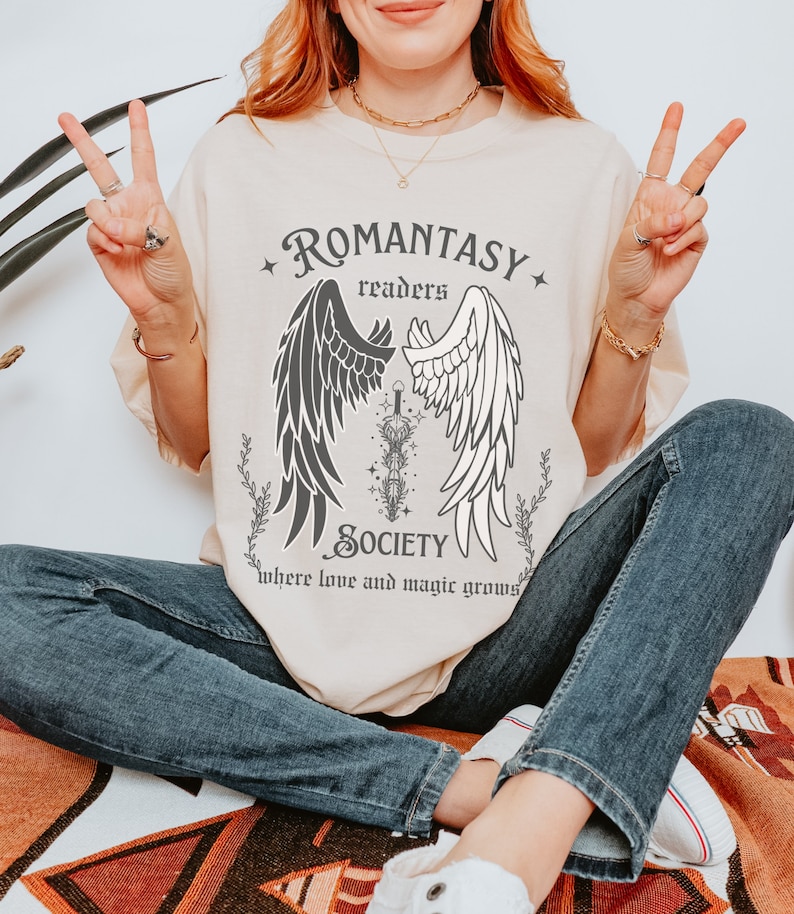 Puede incluir: Una camiseta beige con un dise&ntilde;o gr&aacute;fico de alas de &aacute;ngel blancas y negras con una espada en el medio. El texto "ROMANTASY readers SOCIETY where love and magic grows" est&aacute; impreso en la camiseta.