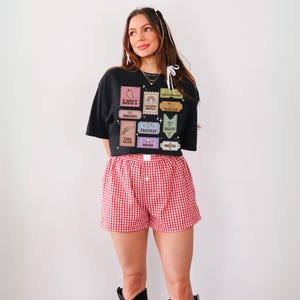 Może przedstawiać: Czarny, kr&oacute;tki t-shirt z nadrukiem kolorowych znaczk&oacute;w przedstawiających r&oacute;żne gatunki książek. Koszulka jest połączona z czerwonymi i białymi szortami w kratę.