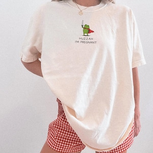 May include: Ivory-colored t-shirt with a cartoon frog graphic holding a sword and wearing a cape. The text "Huzzah I'm Pregnant" is below the frog. The model is wearing red and white checkered shorts.