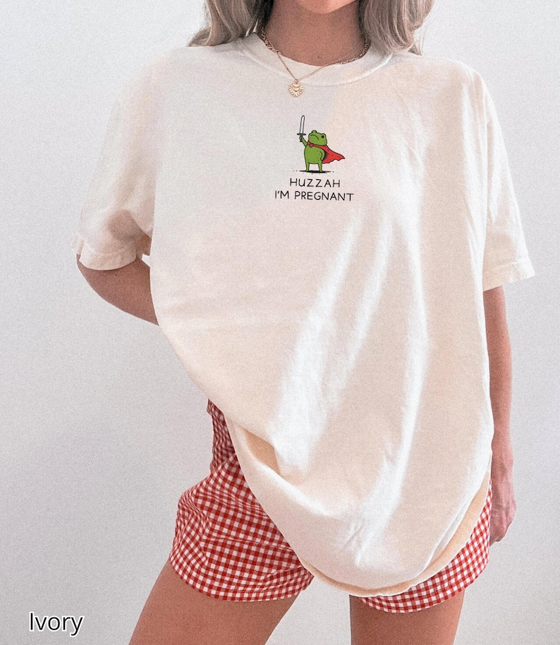 May include: Ivory-colored t-shirt with a cartoon frog graphic holding a sword and wearing a cape. The text "Huzzah I'm Pregnant" is below the frog. The model is wearing red and white checkered shorts.