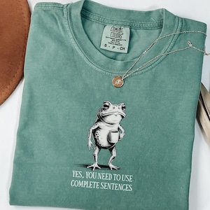 May include: Light green t-shirt featuring a frog graphic holding a mug, with the text "YES, YOU NEED TO USE COMPLETE SENTENCES." A silver necklace with a small pendant is draped across the shirt.