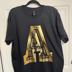 Op de afbeelding: Zwart T-shirt met een groot goudkleurig letter "A" ontwerp. Het ontwerp bevat namen en de nummers "ONE NINE" en "OH SIX". Het shirt hangt aan een zwarte hanger.