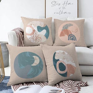 May include: Four square throw pillows in a light beige fabric, each featuring a circular graphic design. The prints include abstract shapes, botanical elements, a crescent moon, and a rainbow in muted tones of blue, terracotta, and cream.