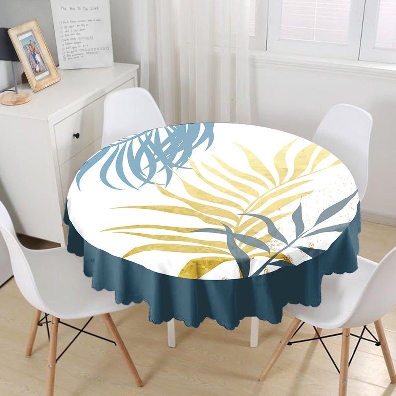 May include: A round tablecloth featuring a white background with a botanical print of large palm leaves in shades of gold and muted blue. The tablecloth has a solid dark blue scalloped edge that drapes over the sides of the circular table.