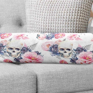 Pode incluir: Uma almofada cilíndrica com fundo branco e um padrão repetitivo de crânios humanos, peónias cor-de-rosa e suculentas azul-escuras. O design inclui delicadas folhas de feto intercaladas entre os elementos florais e os crânios.