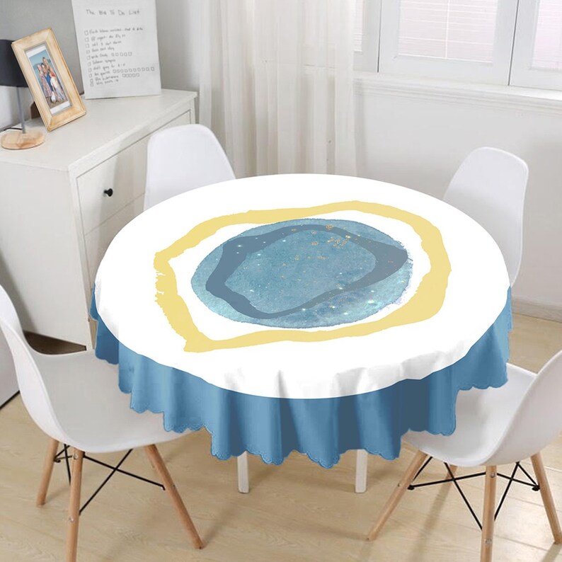 May include: A round tablecloth featuring a white top surface with a central abstract design consisting of a blue textured circle surrounded by a yellow organic border. The tablecloth has a solid blue ruffled skirt with a scalloped edge finish.