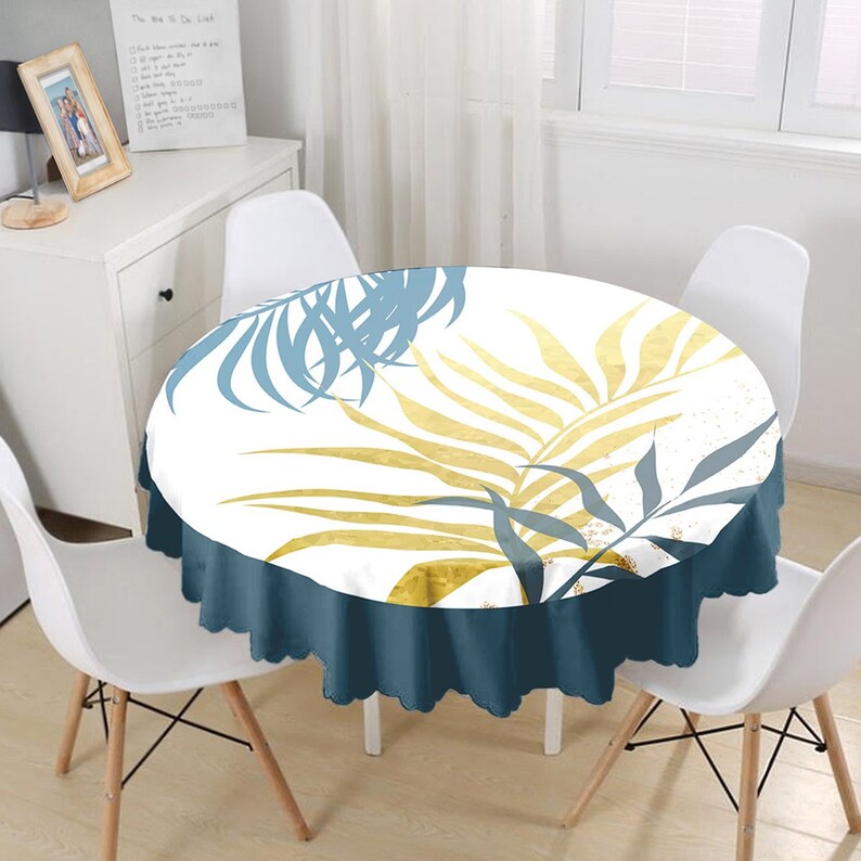 May include: A round tablecloth featuring a white background with a botanical print of large palm leaves in shades of gold and muted blue. The tablecloth has a solid dark blue scalloped edge that drapes over the sides of the circular table.