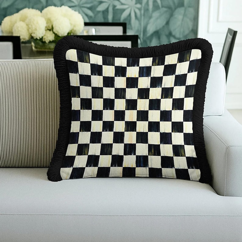Puede incluir: Coj&iacute;n decorativo cuadrado con un patr&oacute;n de cuadros blancos y negros. El coj&iacute;n tiene un borde con flecos negros y est&aacute; colocado sobre un sof&aacute; blanco. El dise&ntilde;o del coj&iacute;n recuerda a un tablero de ajedrez, a&ntilde;adiendo inter&eacute;s gr&aacute;fico.