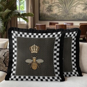 Puede incluir: Dos cojines decorativos con un borde a cuadros blanco y negro y flecos negros. El centro presenta un dise&ntilde;o de abeja y corona bordada en oro sobre un fondo de panal negro.