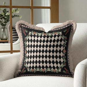 Puede incluir: Coj&iacute;n decorativo cuadrado con un patr&oacute;n central de diamantes negros y crema. El coj&iacute;n tiene un borde negro con dise&ntilde;os florales y de gallos, y una franja beige. Est&aacute; colocado en un sill&oacute;n blanco.