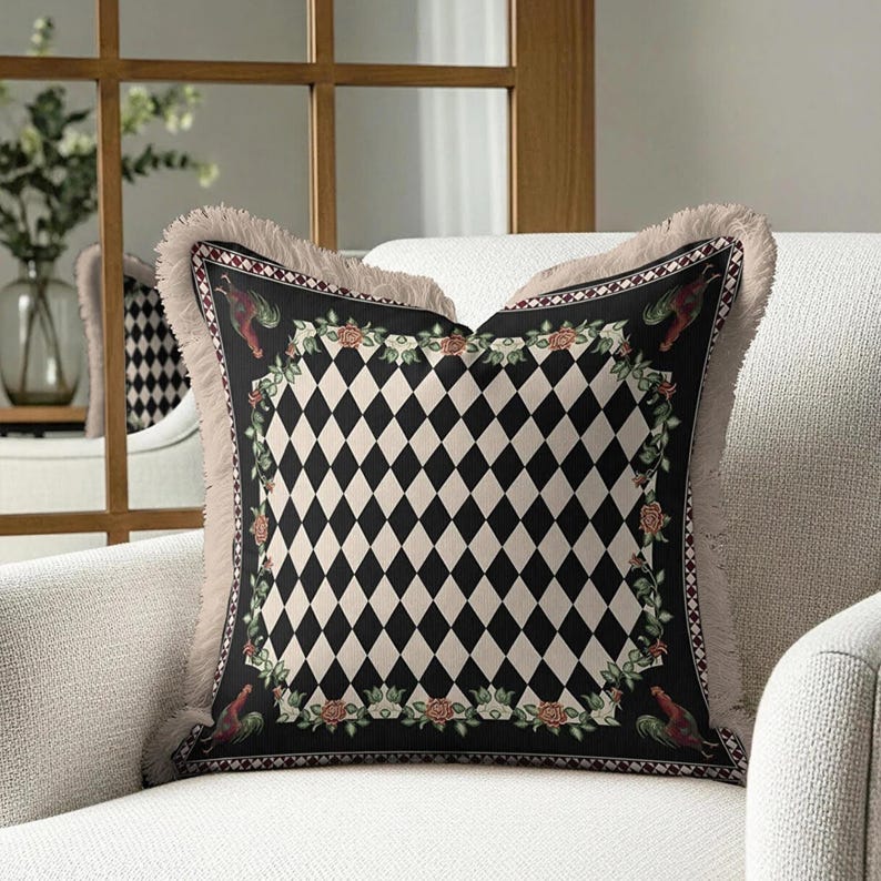 Puede incluir: Coj&iacute;n decorativo cuadrado con un patr&oacute;n central de diamantes negros y crema. El coj&iacute;n tiene un borde negro con dise&ntilde;os florales y de gallos, y una franja beige. Est&aacute; colocado en un sill&oacute;n blanco.
