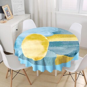 May include: A round tablecloth featuring an abstract watercolor design with a large yellow circle, horizontal yellow bands, and various shades of blue. The fabric has a scalloped edge and drapes over a circular table surrounded by white chairs.