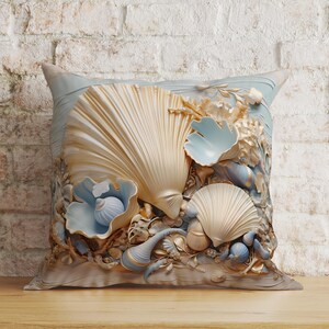 Puede incluir: Un coj&iacute;n cuadrado con un dise&ntilde;o detallado en relieve de varias conchas marinas en tonos crema, dorado y azul p&aacute;lido. La composici&oacute;n central incluye conchas grandes en forma de abanico, conchas en espiral m&aacute;s peque&ntilde;as y texturas de coral.