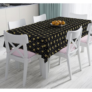 Könnte beinhalten: Eine schwarze Tischdecke mit einem sich wiederholenden Muster aus goldenen Schleifen und weißen Punkten bedeckt einen Esstisch. Weiße Stühle mit rosa Kissen umgeben den Tisch. Ein Teller mit Obst steht auf der Tischdecke.
