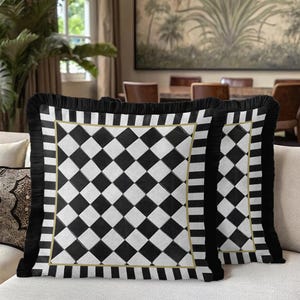 Puede incluir: Dos cojines cuadrados decorativos con un patr&oacute;n de cuadros blancos y negros. Los cojines tienen un borde de flecos negros y un ribete dorado. Se encuentran sobre un sof&aacute; beige, ideales para la decoraci&oacute;n del hogar.