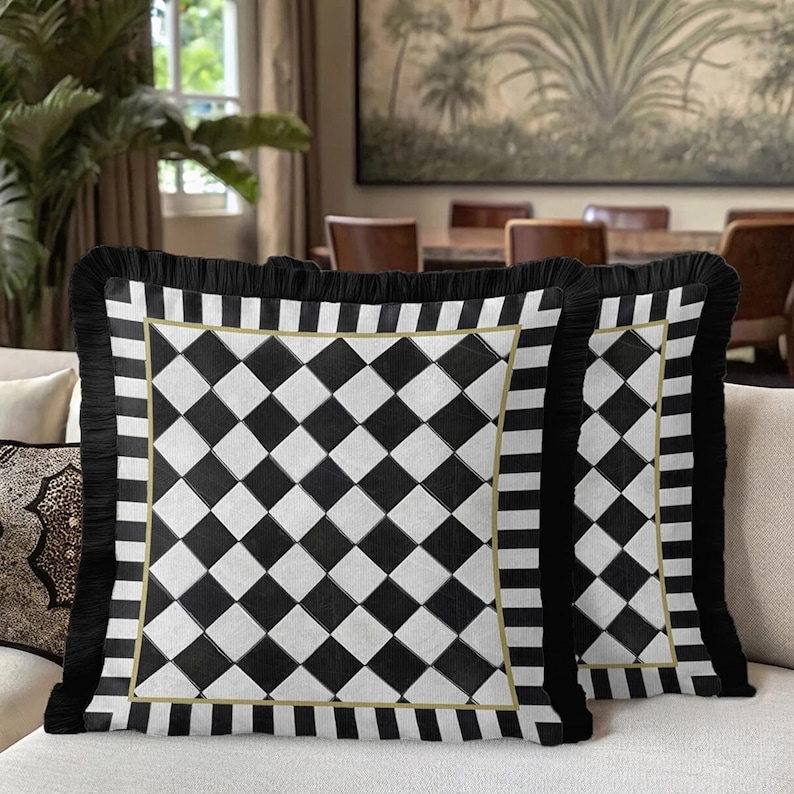 Puede incluir: Dos cojines cuadrados decorativos con un patr&oacute;n de cuadros blancos y negros. Los cojines tienen un borde de flecos negros y un ribete dorado. Se encuentran sobre un sof&aacute; beige, ideales para la decoraci&oacute;n del hogar.