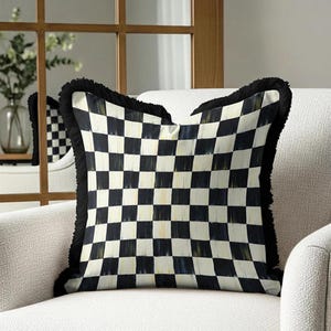 Puede incluir: Un coj&iacute;n decorativo cuadrado con un patr&oacute;n de cuadros blancos y negros. El coj&iacute;n tiene un borde con flecos negros y est&aacute; colocado sobre un sill&oacute;n blanco. El dise&ntilde;o recuerda a un tablero de ajedrez.