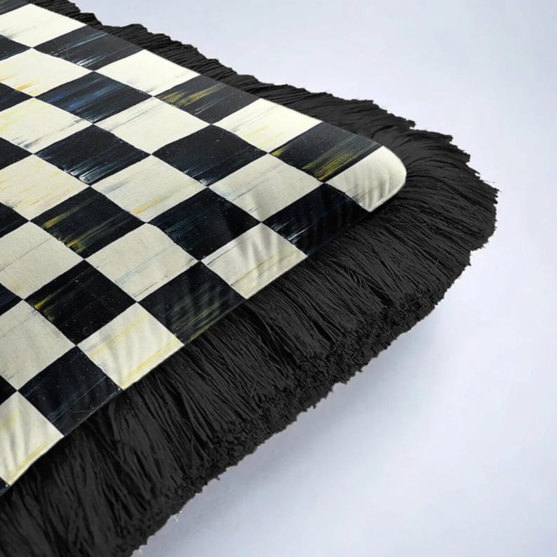 Puede incluir: Un coj&iacute;n decorativo con un patr&oacute;n de cuadros blancos y negros en la parte superior. El coj&iacute;n tiene un grueso borde de flecos negros. El patr&oacute;n de cuadros est&aacute; formado por cuadrados negros y blanquecinos. El coj&iacute;n se utiliza probablemente para la decoraci&oacute;n del hogar.