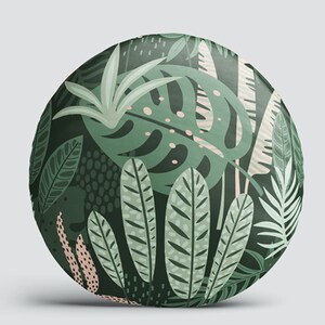 K&ouml;nnte beinhalten: Ein rundes Kissen mit einem tropischen Blattmotiv in Gr&uuml;n und Rosa. Das Kissen ist aus weichem, pl&uuml;schem Stoff gefertigt und zeigt verschiedene Blattformen und -gr&ouml;&szlig;en.