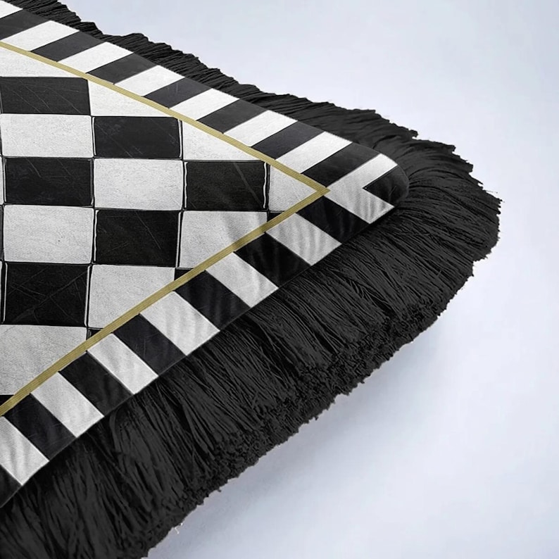 Puede incluir: Un coj&iacute;n decorativo con un patr&oacute;n de cuadros blanco y negro y un borde de flecos negros. El coj&iacute;n tiene un ribete dorado alrededor de una secci&oacute;n triangular con un dise&ntilde;o a rayas.