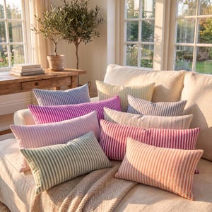 Op de afbeelding: Een collectie rechthoekige sierkussens met verticale strepen in diverse kleuren, waaronder magenta, lichtroze, saliegroen, perzik, geel en grijs, gerangschikt op een lichtgekleurde bank met een textuurrijke plaid eronder gedrapeerd.