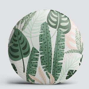 K&ouml;nnte beinhalten: Runder Dekokissen mit einem tropischen Blattmotiv in Gr&uuml;n und Wei&szlig;. Das Kissen zeigt verschiedene Bl&auml;tter, darunter Monstera, Bananen- und Palmenbl&auml;tter.