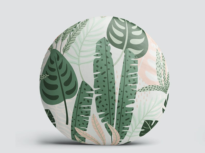 K&ouml;nnte beinhalten: Runder Dekokissen mit einem tropischen Blattmotiv in Gr&uuml;n und Wei&szlig;. Das Kissen zeigt verschiedene Bl&auml;tter, darunter Monstera, Bananen- und Palmenbl&auml;tter.