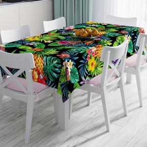May include: A rectangular tablecloth with a vibrant tropical floral pattern featuring pineapples, hibiscus, and palm leaves on a black background. The tablecloth is set on a white table with white chairs and pink cushions.