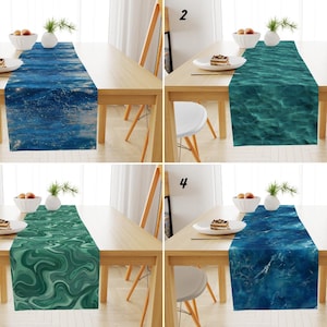 Può includere: Quattro runner da tavolo con disegni a tema oceano sono esposti su tavoli di legno. I runner presentano motivi d'acqua blu e verdi, tra cui onde ed effetti marmorizzati. Ogni tavolo è apparecchiato con un piatto bianco, una ciotola di frutta e un vaso con verde.