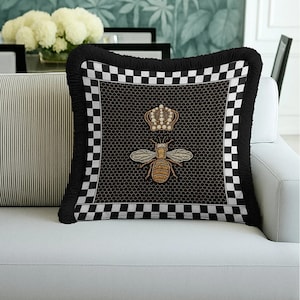 Puede incluir: Coj&iacute;n decorativo negro con dise&ntilde;o de abeja y corona. El coj&iacute;n presenta un fondo negro con un patr&oacute;n de panal, un bordado de abeja y corona, un borde a cuadros y una franja negra.
