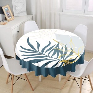 May include: A round tablecloth featuring a white background with a large dark blue palm leaf print, a light gray leaf silhouette, and thin gold line accents. The tablecloth has a solid dark blue scalloped edge trim around the entire perimeter.