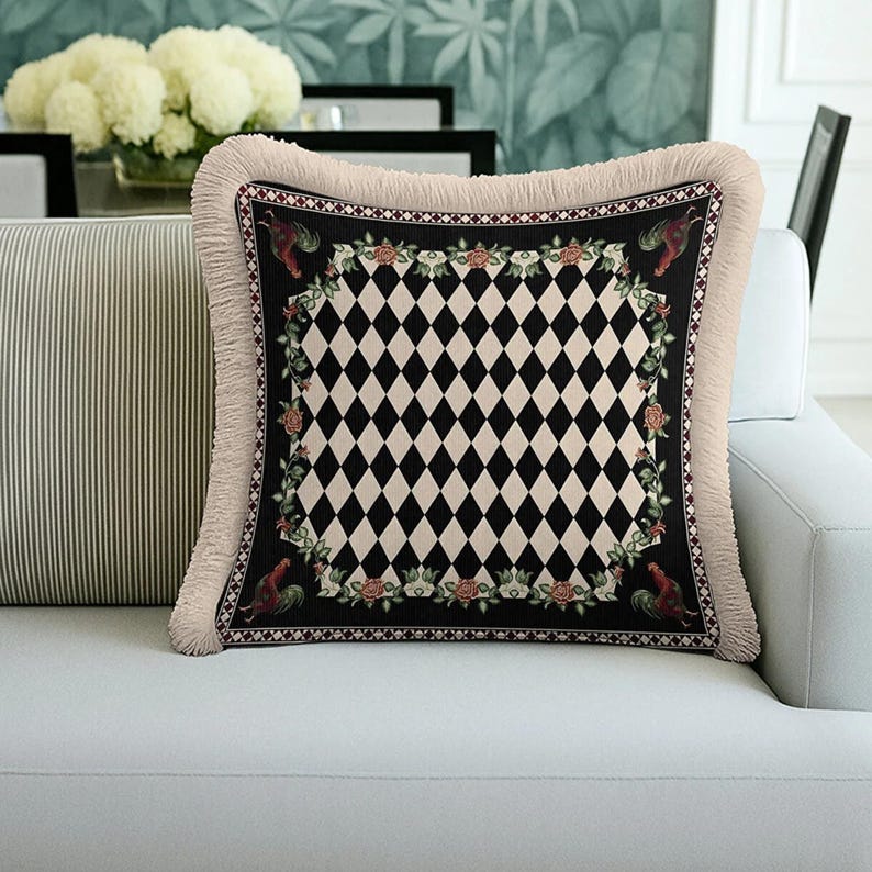 Puede incluir: Coj&iacute;n decorativo cuadrado con un patr&oacute;n central de diamantes en blanco y negro. Tiene un borde negro con dise&ntilde;os florales y de gallos, y un fleco beige. El coj&iacute;n est&aacute; sobre un sof&aacute; blanco.