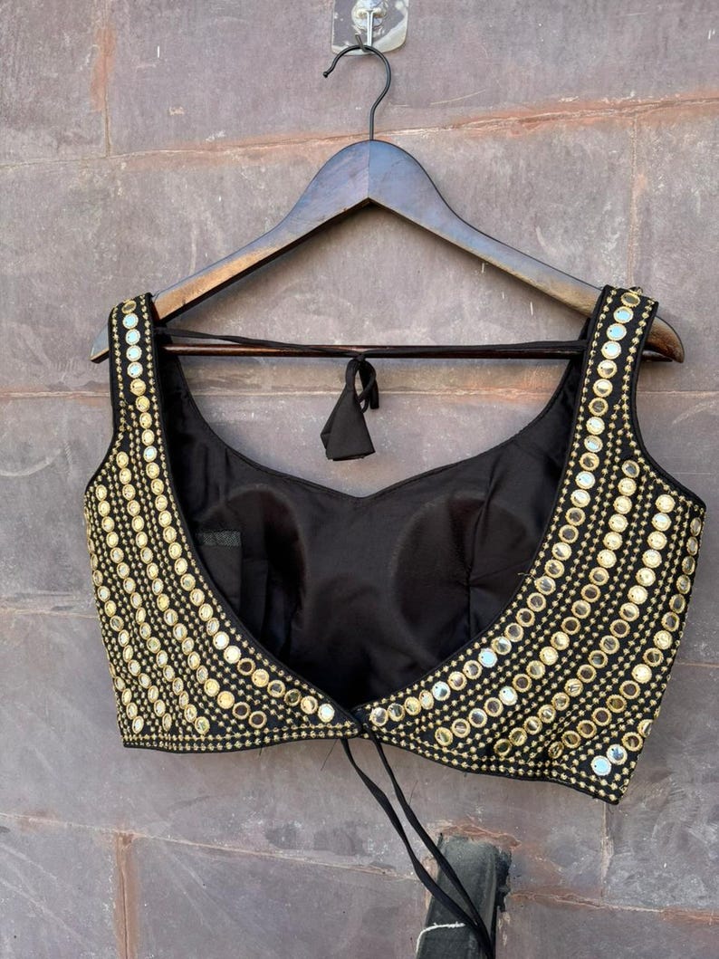 May include: A black sleeveless blouse featuring a wide scoop neckline and a cropped silhouette. The front panels are heavily embellished with rows of small, circular gold-toned mirrors and metallic gold embroidery, creating a dense, reflective pattern.