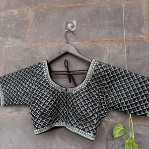 May include: Black blouse with a geometric pattern and silver-colored trim. The blouse has short sleeves and is hanging on a wooden hanger. The fabric has a diamond-shaped design.