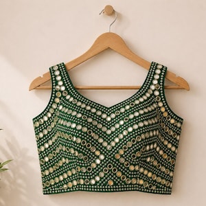 May include: A sleeveless dark green blouse featuring a V-neckline and an intricate pattern of small, circular reflective mirrors arranged in horizontal and chevron-like rows across the entire front surface, suspended on a wooden hanger.