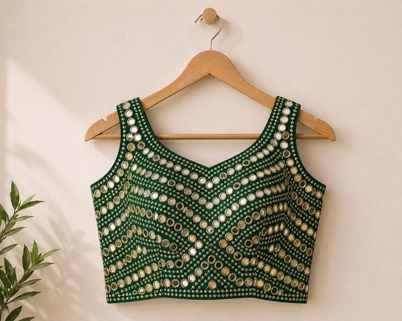 May include: A sleeveless dark green blouse featuring a V-neckline and an intricate pattern of small, circular reflective mirrors arranged in horizontal and chevron-like rows across the entire front surface, suspended on a wooden hanger.