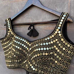 May include: A black sleeveless blouse featuring a scoop neckline and intricate gold-toned embroidery. The garment is densely decorated with numerous small, circular reflective mirrors arranged in curved, repeating patterns across the entire front surface.