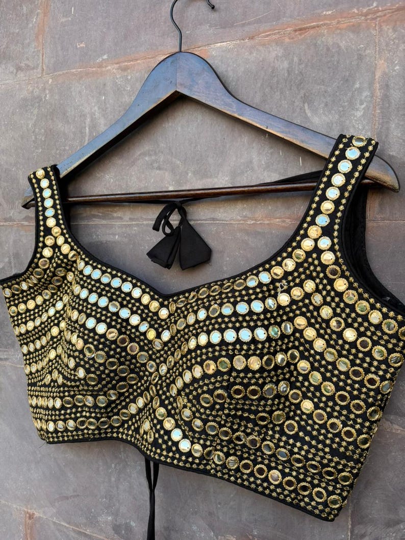 May include: A black sleeveless blouse featuring a scoop neckline and intricate gold-toned embroidery. The garment is densely decorated with numerous small, circular reflective mirrors arranged in curved, repeating patterns across the entire front surface.