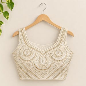 Puede incluir: Una blusa sin mangas de color crema con escote redondo y bordados intrincados. La prenda está adornada con hilo metálico dorado, pequeños espejos circulares y cuentas blancas dispuestas en un patrón simétrico de festones.
