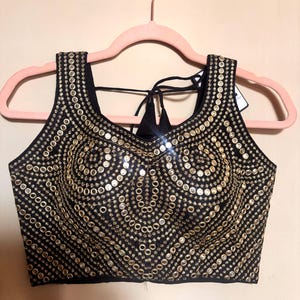 May include: Black sleeveless top embellished with gold sequins arranged in a circular design. The top is displayed on a pink hanger. The sequins provide a shimmering appearance, suitable for events. The garment features a tie-back.