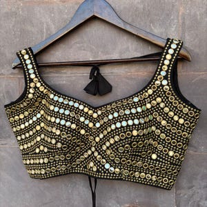 May include: Black sleeveless top with gold circular embellishments arranged in wavy lines. The top has a V-neckline and is hanging on a wooden hanger. A black tie is visible at the back of the top.