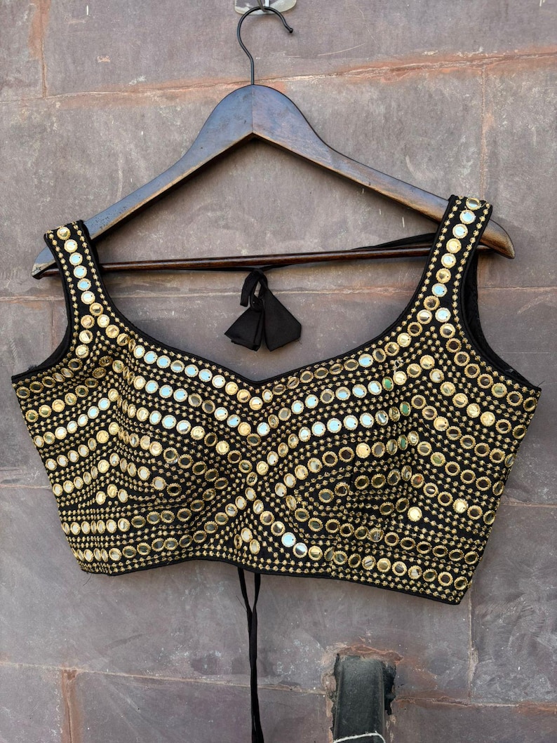 May include: A black sleeveless blouse featuring a sweetheart neckline and a dense pattern of circular gold-toned mirrors and metallic embroidery. The garment includes a tie-back closure with black fabric tassels hanging from the centre back.
