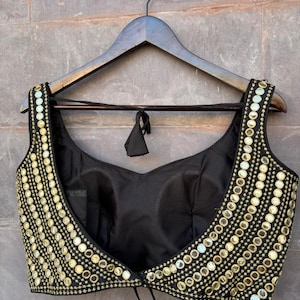 May include: A sleeveless black blouse featuring a deep scoop neckline and wide shoulder straps. The garment is heavily embellished with rows of small, circular gold-toned mirrors and intricate gold embroidery along the front edges and straps.