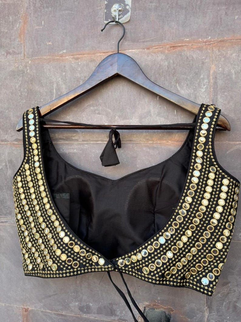 May include: A sleeveless black blouse featuring a deep scoop neckline and wide shoulder straps. The garment is heavily embellished with rows of small, circular gold-toned mirrors and intricate gold embroidery along the front edges and straps.