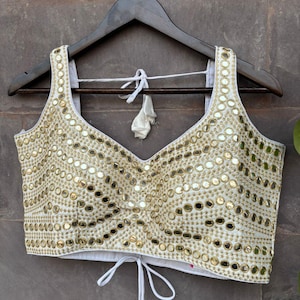 May include: Ivory-colored, sleeveless top with a V-neck and intricate gold mirror embellishments. The top is hanging on a wooden hanger. The bottom of the top has a drawstring closure with a white tassel.