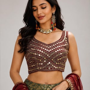 May include: A sleeveless maroon blouse featuring a V-neckline and intricate gold embroidery with circular mirror embellishments arranged in a geometric pattern. The garment is paired with a dark green skirt and a matching maroon sheer dupatta.