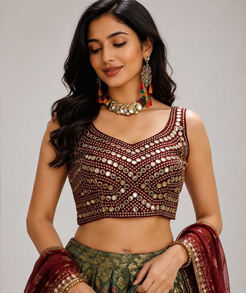 May include: A sleeveless maroon blouse featuring a V-neckline and intricate gold embroidery with circular mirror embellishments arranged in a geometric pattern. The garment is paired with a dark green skirt and a matching maroon sheer dupatta.