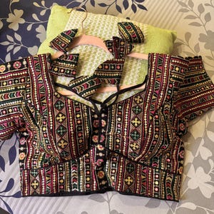 May include: An embroidered black blouse with a decorative pattern in red, green, and gold. The blouse has short sleeves and a lace-up front. Matching embroidered accessories are also visible.