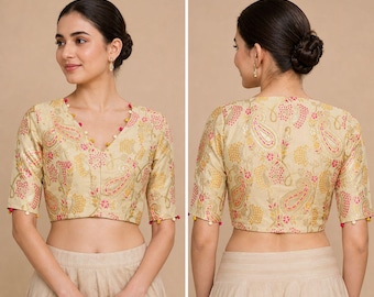 May include: A beige crop top with a V-neck, featuring a pink and gold paisley design. The short sleeves and neckline are trimmed with small, colourful beads. This blouse is a component of a traditional Indian outfit.