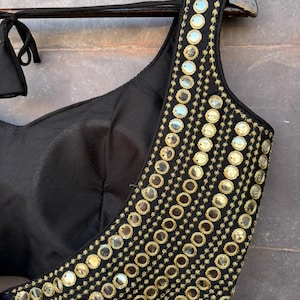 May include: A black sleeveless blouse featuring a wide shoulder strap adorned with multiple rows of circular reflective mirrors and gold-toned embroidery. The garment has a sweetheart neckline and includes thin black fabric ties at the shoulder.